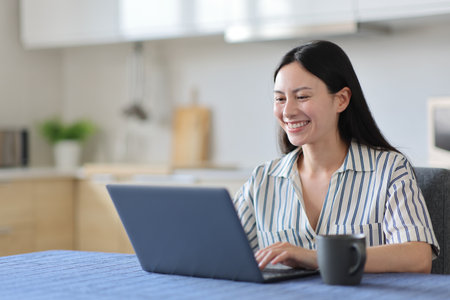 Happy asian woman using laptop in the kitchen at homeの写真素材