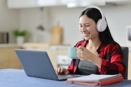 Happy asian student using headphone and laptop e-learning in the kitchen at homeの写真素材