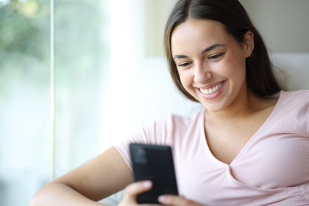 Happy woman using cell phone smiling sitting on a couch at homeの写真素材