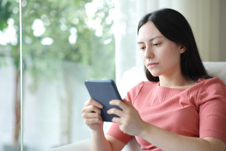 Serious asian woman reading ebook on a sofa at homeの写真素材