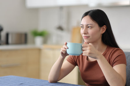 Relaxed asian woman drinking and contemplating in the kitchen at homeの写真素材