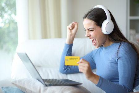 Excited woman wearing headphone and using laptop buying music or videos online with credit card sitting on a couch at homeの写真素材