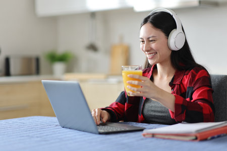 Happy asian student e-learning with headphone and laptop at breakfast drinking orange juice at homeの写真素材