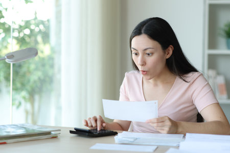 Surprised asian woman calculating expenses holding bank statement at homeの写真素材