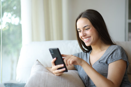 Happy woman enjoying using her smart phone sitting on a couch at homeの写真素材