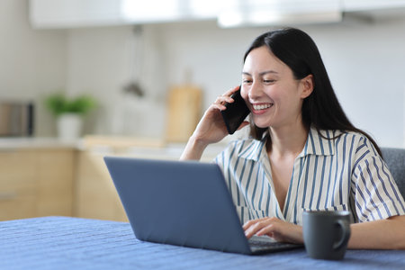 Happy asian woman talking on phone checking laptop in the kitchen at homeの写真素材