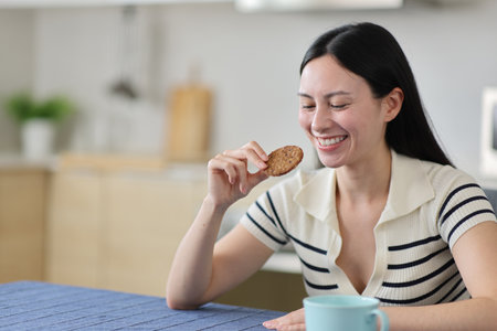Happy asian woman eating cookie in the kitchen at homeの写真素材