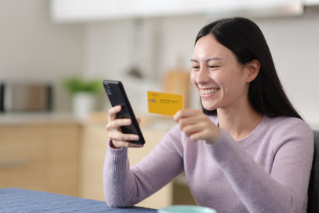 Happy asian woman buying online using card and phone in the kitchen at homeの写真素材