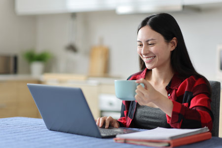 Happy asian woman drinking and e-learning using laptop in the kitchen at homeの写真素材