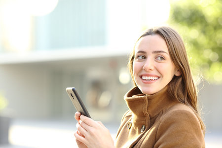 Woman holding phone in winter with perfect smile in the street looking at sideの写真素材