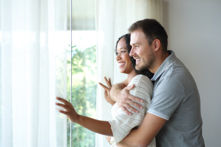 Happy interracial couple at home contemplating outside through the windowの写真素材