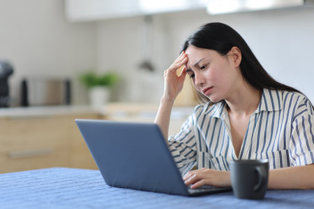 Worried asian woman checking laptop complaining in the kitchen at homeの写真素材
