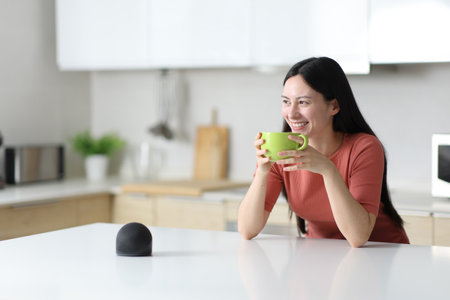 Happy asian woman drinking in the kitchen at home with smart speaker