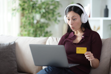 Surprised asian woman wearing headphone buying online media with credit card and laptop sitting on a sofa at homeの写真素材
