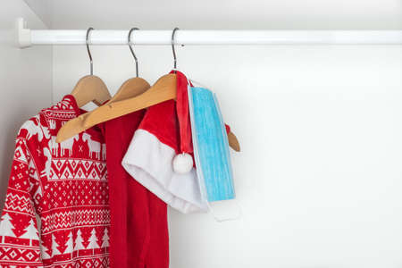 Horizontal color image with a front view of a white and red christmas pajamas, hat, tights and face mask hanging on wooden hangers inside a white closet. Holidays during coronavirus pandemic.の写真素材