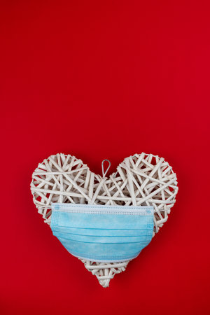 Vertical color image with a front view of a white heart with a blue face mask on one side of a red background. Concept of a Valentines day during Coronavirus or Covid pandemic.の写真素材