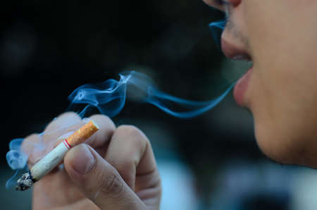 Young man smoking cigarette on black background with smokeの写真素材