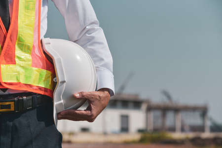 Construction foreman holding white hat safety hard hat on building backgroundの写真素材