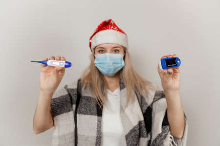 Woman in santa hat, wrap and protective medical mask holds thermometer and puls oximetrの写真素材