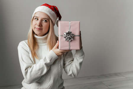 Woman in a white sweater and a red santa hat holding a pink gift in her hands on a gray background