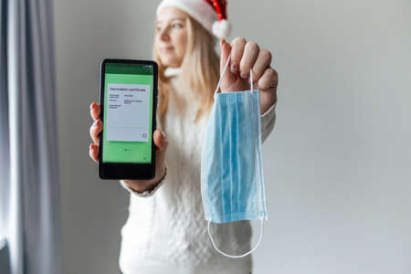Woman in santa hat holds phone with vaccination certificate and protective medical mask
