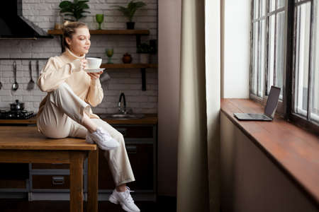 Happy young woman is in a cozy home atmosphere, sits at the kitchen table and holds a cup of coffee...