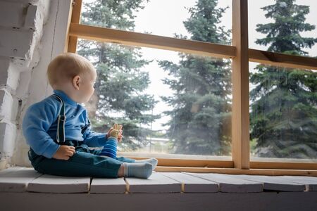 Beautiful blue-eyed baby sitting on the windowsill and looks out the window at the green trees.の写真素材