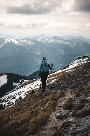 A girl in a blue-gray jacket, dark-gray hat, black leggings, blue-green backpack, and trekking poles...