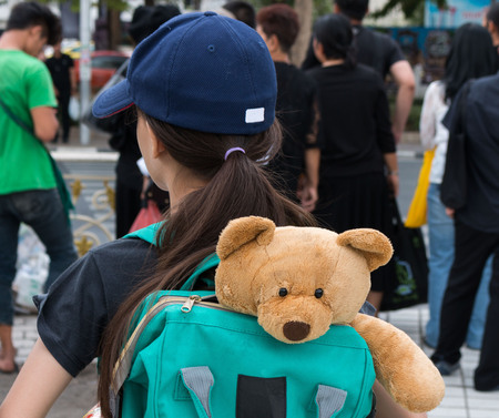 A young girl with a stuffed animal teddy bear in the bag on her back. Cute bear stay in bag on young girl. A girl moving pass people go to her home feeling like lonely.の写真素材