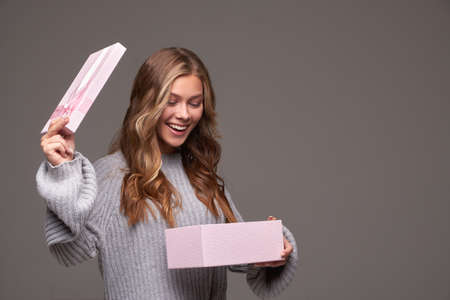 Portrait of a surprised girl holding a pink gift box in gray clothes on a gray background