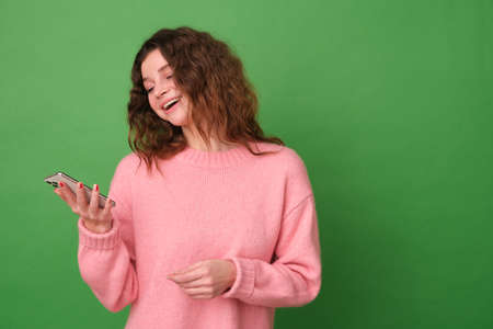 A young happy girl is smiling in a pink sweater and holding a phone on a green backgroundの写真素材