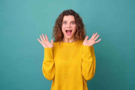 Amused cute girl with curly hair, raises her palms, has a cheerful expression, smiles widely, sees something funny, wears a yellow sweater isolated on a blue backgroundの写真素材