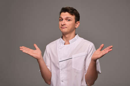 A young doctor on a gray background in white clothes shrugs and does not know what to doの写真素材