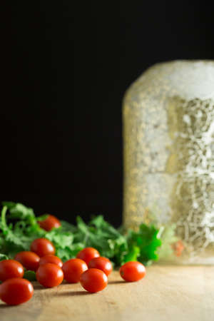 Cherry tomatoes and baby kale next to glass object on wooden cutting board with black backgroundの写真素材