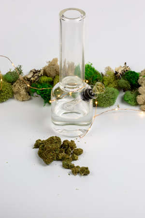 Glass bong surrounded by weed buds, string lights and moss against white backgroundの写真素材
