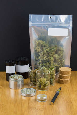 assorted medical marijuana in containers and plastic bags with white labels, grinder, vape pen and kief on bamboo table with black background in portraitの写真素材