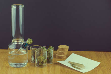 Medical marijuana in glass containers next to glass bong with cone rolling paper angled to the right on bamboo table and black backgroundの写真素材