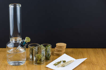 Medical marijuana in glass containers next to glass bong with cone rolling paper angled to the left on bamboo table and black backgroundの写真素材