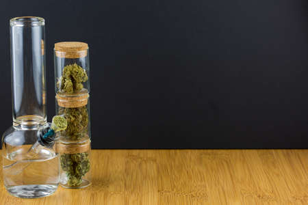 Stacked glass containers filled with assorted medical marijuana next to glass bong on bamboo surface with black backgroundの写真素材