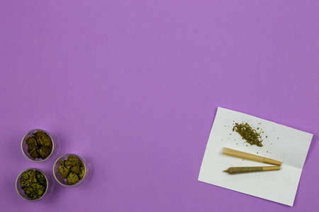 Marijuana in glass containers with cone shaped rolling paper and joint on white paper against ultraviolet backgroundの写真素材