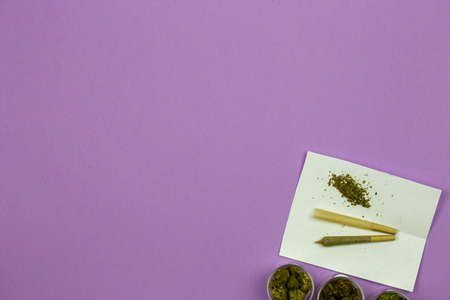 Marijuana in glass containers with cone shaped rolling paper and joint on white paper in bottom right corner against ultraviolet backgroundの写真素材