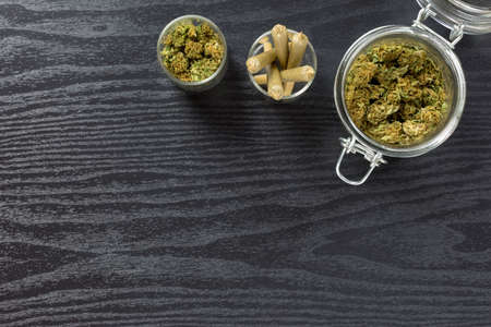 Assorted weed buds and cone shaped joints in mixed sized glass jars on black textured surfaceの写真素材