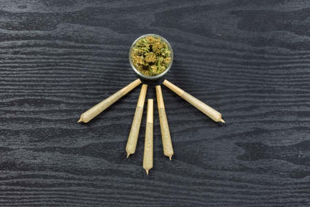 Weed buds in small glass jar with cone shaped joints arranged on black textured surfaceの写真素材