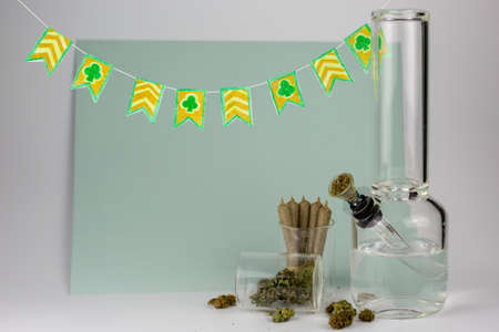 Weed buds in glass jar with cone joints, packed bong and hand crafted St. Patricks day pennants against a blue and white backdropの写真素材