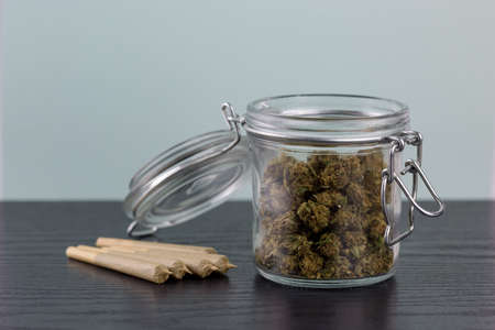 Marijuana in glass jar with metal clasp, next to joints arranged on black wood textured surface with a white backdropの写真素材