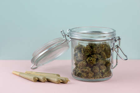 Marijuana in glass jar with metal clasp, next to brown cone joints arranged on pastel pink and blue backdropの写真素材