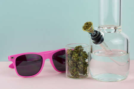 Packed clear glass bong with weed buds in a small glass jar and pink sunglasses on a blue and pink pastel backdropの写真素材