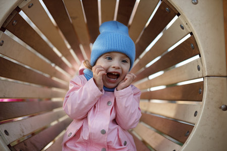 Cute little girl in a pink jacket and blue hat on a wooden swing.の写真素材