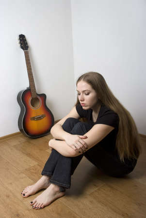 Sad woman with long brown hair sitting on the wooden floor with her acoustic guitar in the background.の写真素材