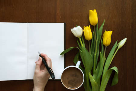 Female hand writing in a book next to a cup of coffee and tulips on a wooden table.の写真素材